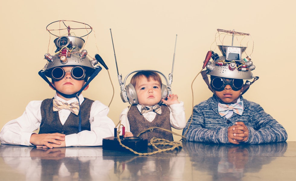 A young boy imagines reading minds of his two friends with a homemade science project. They are dressed in casual clothing, glasses and bow ties. They are serious and sitting at a table with helmets on their heads in front of a beige background. To illustrate article about average IQ of lawyers