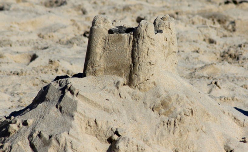 Photo showing a mound of sand at the beach, which has a square sandcastle standing at the top and another at the side. The sandcastles have been made with a moulded bucket and spade, which features turrets on the corners.