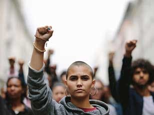 Young woman protesting on the street with her fist raised in air. Group of protesters on the road with their arms raised.